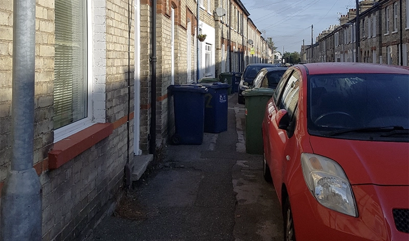 Narrow street with wheelie bins blocking the path and cars parked over the kerb