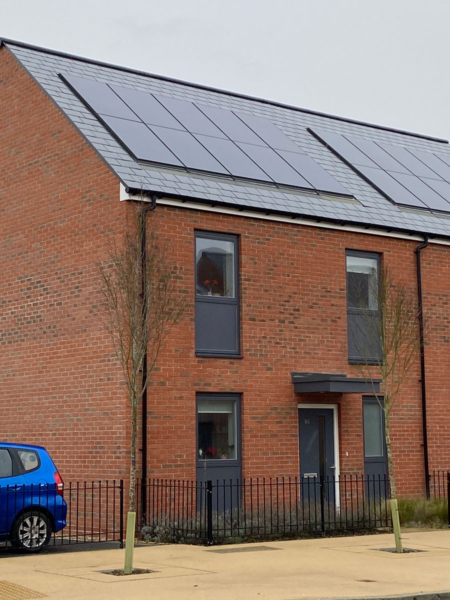Ecohome in Bicester, features solar panels on the roof, a small planted area in fromt of the house and a traditional four windows to the front with a door in the middle.