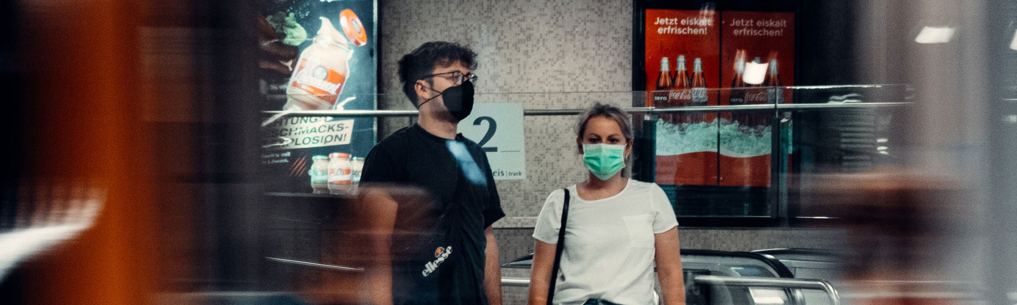 Two people wearing face coverings, standing in front of an escalator